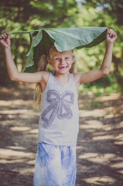 Pequeña niña encantadora sosteniendo una bardana verde sobre su cabeza y sonriendo felizmente — Foto de Stock