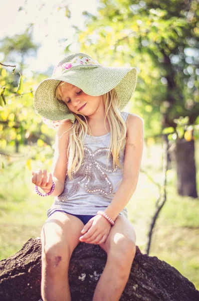 Kleines süßes blondes Mädchen mit Hut sitzt im Baumstamm — Stockfoto