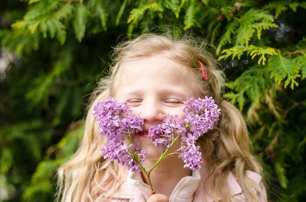 ライラック色の花を押しながら咲く薄紫色のブッシュの横にある屋外に臭いがするかわいいブロンドの女の子 — ストック写真