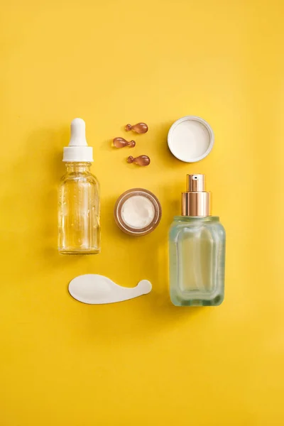 Cilt tazeleme konsepti - cam yüz serumu şişesi, göz kremi, sarı arka planda yüz yağı kapsülleri.
