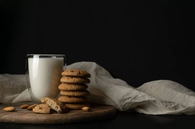 Oat cookies with chocolate, almond, milk is poured into a glass on white cloth over black background. Still life