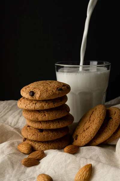 Oat cookies with chocolate, almond, milk is poured into a glass on white cloth over black background — Stockfoto