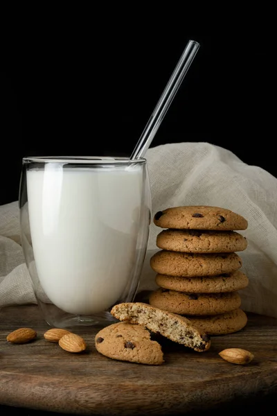 Haver koekjes met chocolade, amandel, melk in glas met dubbele wanden, glazen buis op houten plank over witte doek — Stockfoto