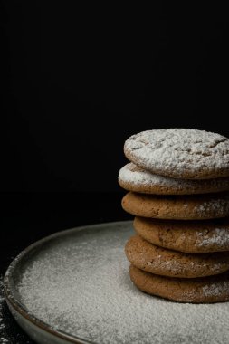 Bir yığın yulaf ezmeli kurabiye, bir parça çikolata ve siyah zemin üzerindeki kil tabakta pudra şekeri. Yakın plan.