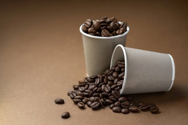 Дві сірі паперові чашки, наповнені кавовими зернами на коричневій поверхні . — стокове фото