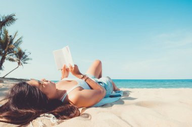 Eğlence yaz - genç kadın tropikal bir kumsalda yatıp kitapla rahatlayın. Mavi deniz içinde belgili tanımlık geçmiş. Yaz tatil kavramı. Vintage renk tonu.
