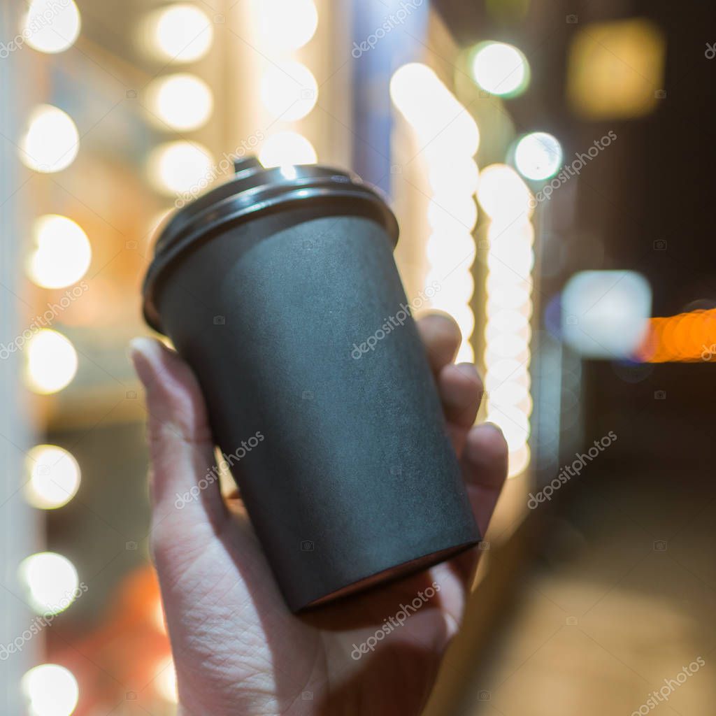 Paper cup of coffee in hand on a background of beautiful bokeh of city lighting. Place for text logo.