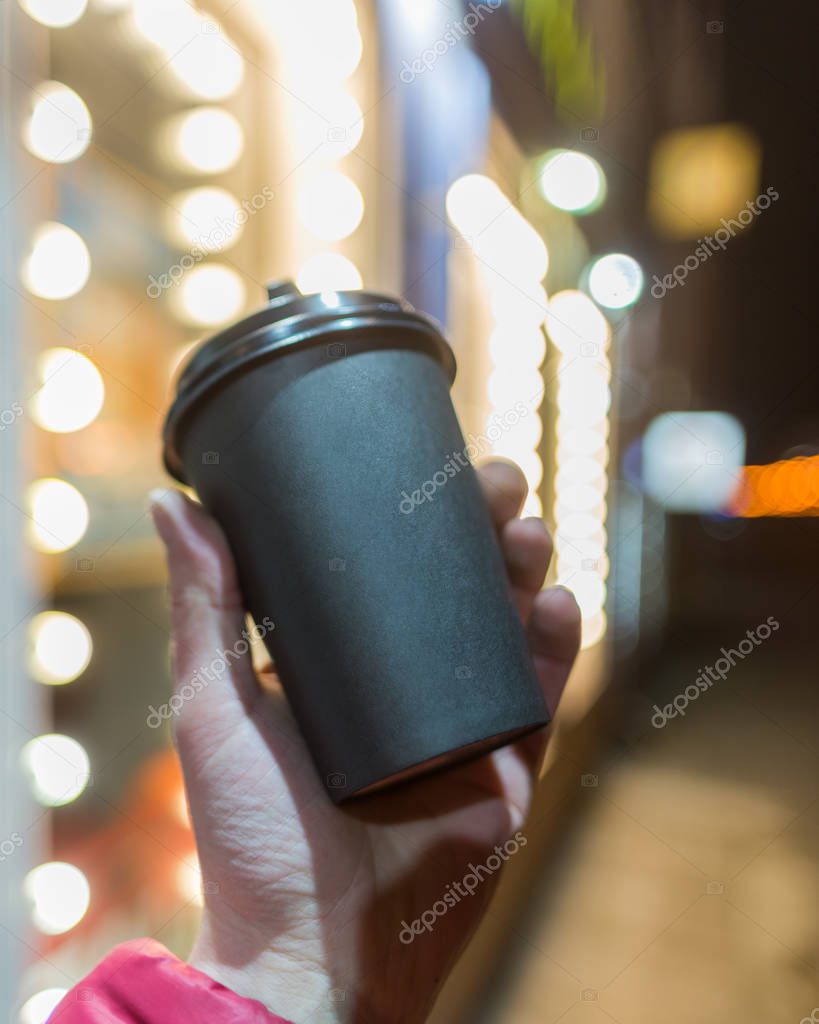 Coffee paper cup in hand on a background of beautiful bokeh of city lighting. Place for text logo.