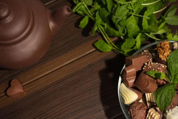 Brown clay teapot, mint, chocolate candies and cinnamon on a brown wooden background. Top view.