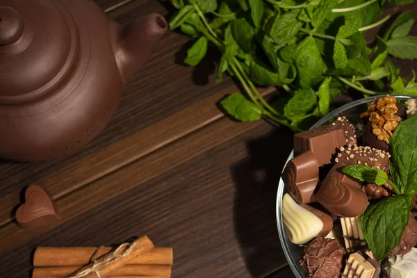 Brown clay teapot, mint, chocolate candies and cinnamon on a brown wooden background. Top view.