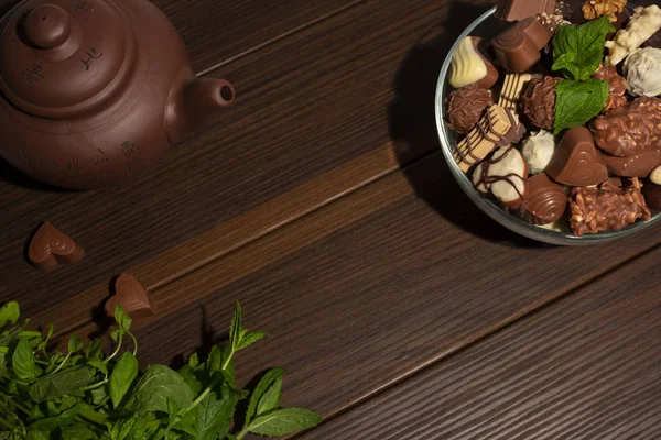 Brown clay teapot, mint, chocolate candies and cinnamon on a brown wooden background. Top view.