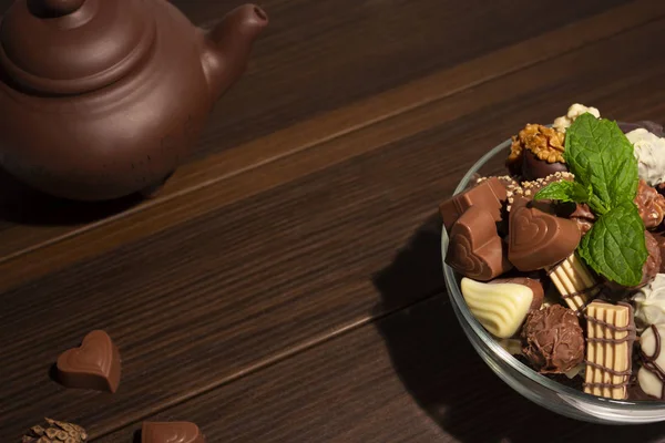 Brown clay teapot, mint, chocolate candies and cinnamon on a brown wooden background. Top view.