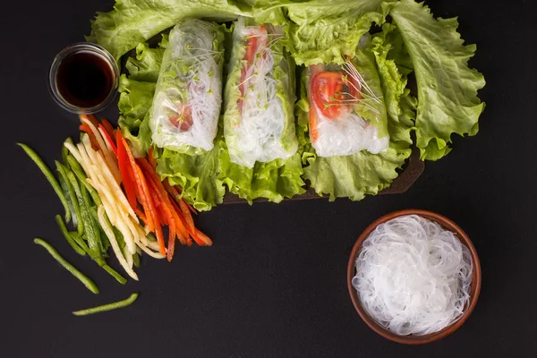 Jarní závitky se zeleninou na černém pozadí. Vedle ingrediencí jsou nakrájené papriky, nudle a sójová omáčka. Vegetariánské jídlo — Stock fotografie