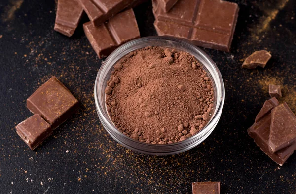 Cacao en polvo en un plato sobre un fondo negro junto al chocolate. primer plano —  Fotos de Stock