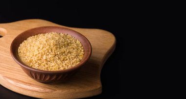 Siyah arka planda, tahta bir tahtanın üzerindeki bir kasede çiğ bulgur. Boşluğu kopyala