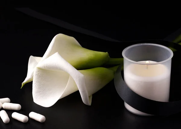 A candle with a mourning black ribbon next to a white flower and pills. The concept of mourning and loss as a result of coronavirus