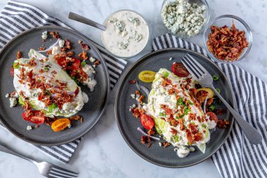 Mavi peynirli salata soslu iki buzdağı marul salatası, pastırma, domates, soğan, yukarıdan atılmış gri taş kaplar üzerinde duran soğanlar. Etrafı malzemelerle çevrili. Keto.