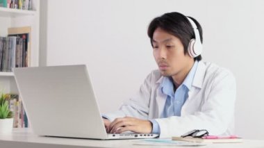 Asyalı erkek doktor kulaklık takıyor ve dizüstü bilgisayarla çalışan beyaz önlük giyiyor. Asyalı doktor dizüstü bilgisayarı ciddi duygularla izliyor