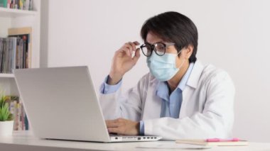 Asyalı Erkek Doktor Maske ve Beyaz Ceket Giyer veya Önlük ve Dizüstü bilgisayarla çalışan Gözlükleri Ayarla. Asyalı doktor dizüstü bilgisayarı ciddi duygularla izliyor