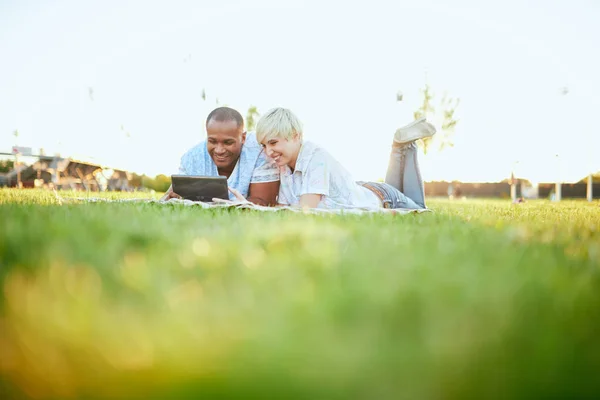Pareja de carrera mixta de millennial en un campo de hierba mirando una tableta digital y leyendo para su próximo periódico escolar — Foto de Stock
