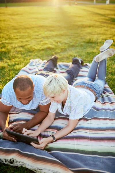 Mixed race couple of millennial in a grass field looking at a digital tablet and reading for their next school paper — Stock Photo, Image