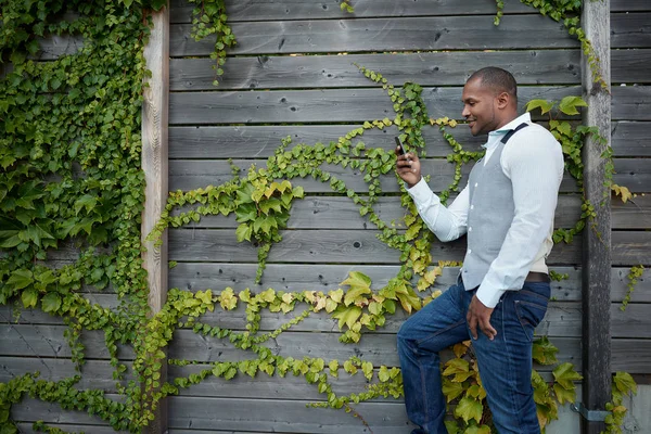 Handsome black man hanging out and texting with his mobile smartphone outside next to a wooden fence