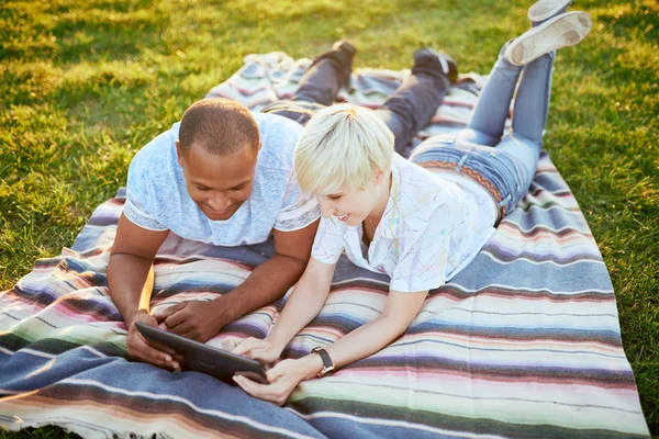 Mixed race couple of millennial in a grass field looking at a digital tablet and reading for their next school paper — Stock Photo, Image