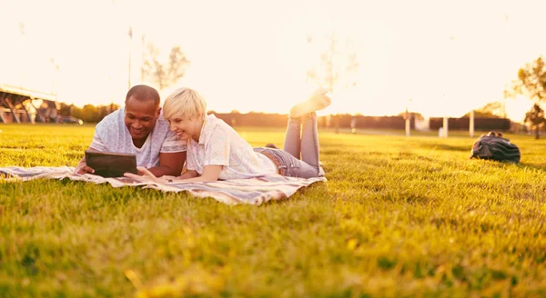 Coppia di razza mista di millennial in un campo di erba guardando un tablet digitale e leggendo per il loro prossimo giornale scolastico — Foto Stock