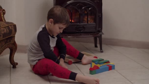 Niño autista en pijama de Navidad sentado en el suelo junto a la chimenea y jugando con bloques de madera en cámara lenta . — Vídeos de Stock