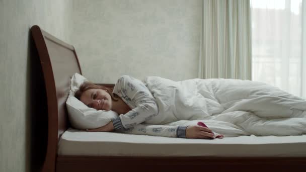 Jonge glimlachende brunette vrouw in heldere pyjama en slaapmasker ontwaken in de slaapkamer in slow motion. Vrouwen nemen de tijd genieten van de ochtend in bed. — Stockvideo