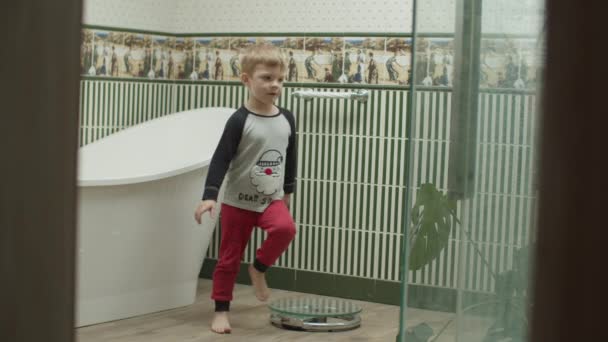 Blondine jongen in pyjama spelen in de badkamer met vloerschalen voor gewichtsbeheersing. Kind staat op glazen weegschaal als moeder. — Stockvideo