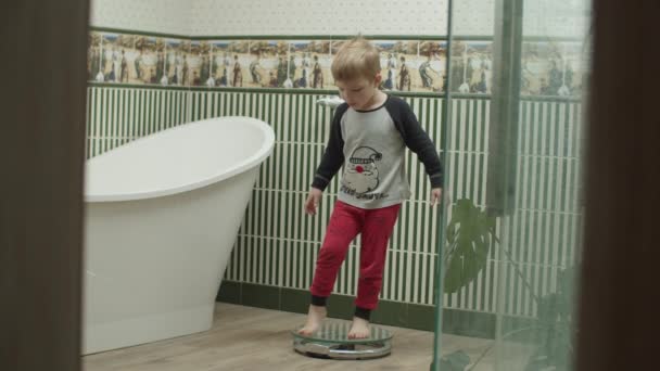 Blonder Junge im Schlafanzug spielt im Badezimmer mit Bodenwaage zur Gewichtskontrolle. Kind steht wie Mutter auf Glaswaage. — Stockvideo
