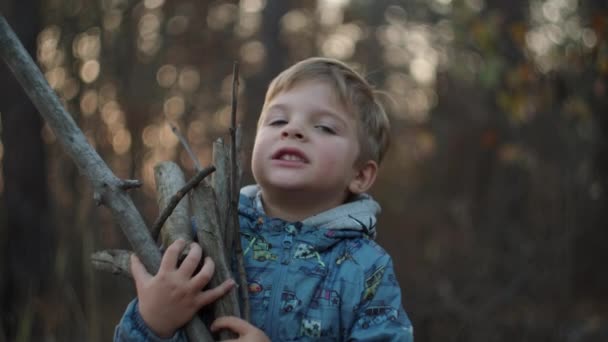 Mittelplan eines Jungen mit trockenen Ästen im Herbstwald bei Sonnenuntergang in Zeitlupe. Blondes Kind lächelt mit Ästen in der Hand in die Kamera. — Stockvideo