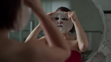 Duştan sonra kırmızı havluyla genç bir kadın aynanın önünde duruyor ve yüzüne ağır çekimde maske takıyor. Islak saçlı kadınların yansıması.. 