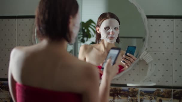 Giovane femmina in maschera cosmetica sul viso dopo la doccia in piedi davanti allo specchio e navigare online in smartphone al rallentatore. Riflessione delle donne con capelli bagnati simpatia foto online . — Video Stock