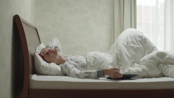 Молодая брюнетка в яркой пижаме и маске для сна просыпается от будильника на планшетном компьютере в спальне в замедленной съемке . — стоковое видео