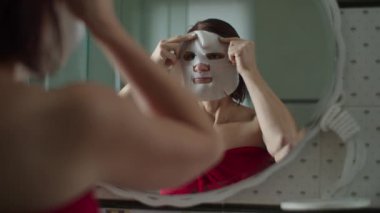 Duştan sonra kırmızı havluyla genç bir kadın aynanın önünde duruyor ve yüzüne ağır çekimde maske takıyor. Islak saçlı kadınların yansıması.. 