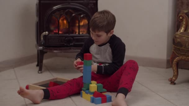 Garçon autiste en pyjama de Noël assis sur le sol près de la cheminée et jouant avec des blocs de bois au ralenti . — Video