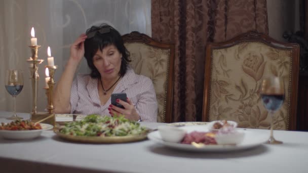 Brunette femmes adultes assises à la table du dîner et surfant en ligne sur smartphone en attendant la fille pour déjeuner avec du vin au ralenti . — Video