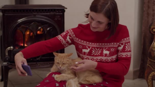 Młoda kobieta w czerwonej piżamie świątecznej siedząca na podłodze z czerwonym kotem na nogach przy kominku i robiąca zdjęcia na smartfonie w zwolnionym tempie — Wideo stockowe