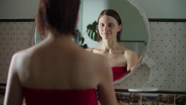 Young female in red towel after shower standing in front of the mirror and applying moisturizing cream on her face in slow motion. Reflection of women with wet hair. — Stockvideo