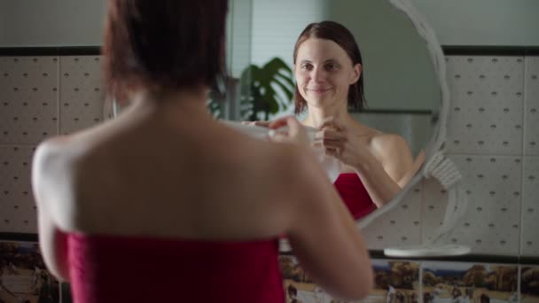 鏡の前に立ち、ゆっくりとした動きで顔にマスクを塗ると、赤いタオルに身を包んだ若い女性。濡れた髪の女性の反射. — ストック動画