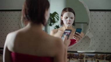 Kozmetik maskeli genç bir kadın duştan sonra aynanın önünde duruyor ve akıllı telefondan yavaş çekimde sörf yapıyor. İnternette ıslak saçlı kadınların resimlerden hoşlanmasının yansıması.. 