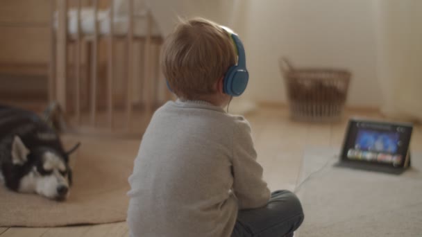 ヘッドフォンでブロンドの男の子が床に座って、犬が近くに横たわっているタブレットコンピュータ上で漫画をオンラインで見ています。シベリアハスキーと子供たち. — ストック動画