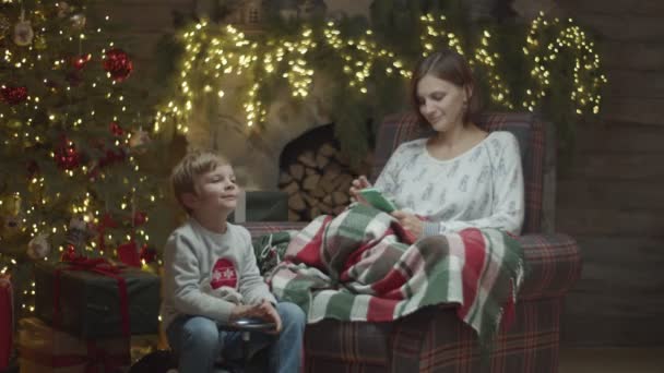 Young female in pajamas in armchair surfing online on smartphone with blonde son playing on toy car in front in Christmas decorations in slow motion. — Stock Video