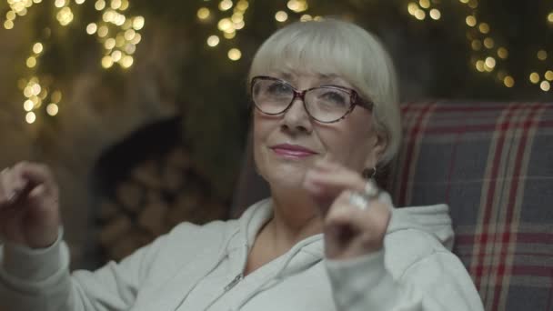 Femme blonde senior dans un fauteuil enlevant des lunettes dans les décorations de Noël avec des lumières floues derrière au ralenti . — Video