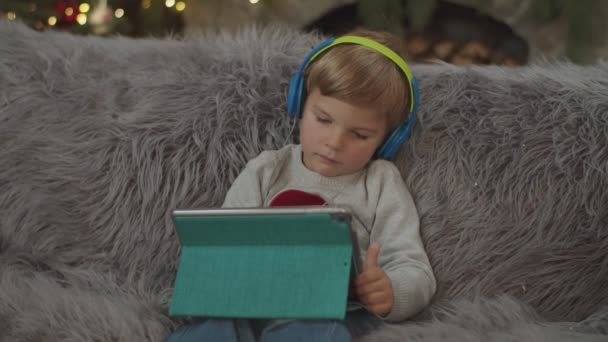 Baby boy with headphones playing online on tablet computer in Christmas decoration. Kid on sofa with gadget in slow motion. — Stock Video