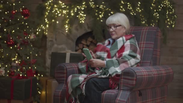 Abuela rubia en manta sentada en sillón junto al árbol de Navidad y surfeando en línea en gadget. Mujeres mayores con smartphone . — Vídeo de stock