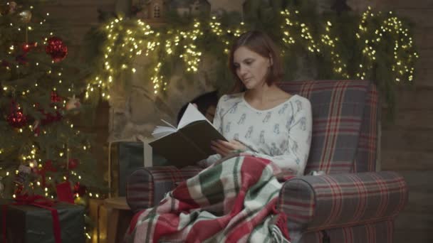 Mujer morena leyendo libro de papel sentado en sillón acogedor con con árbol de Navidad y luces borrosas en la chimenea detrás en cámara lenta . — Vídeos de Stock
