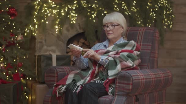 Seniorin in Decke sitzt im Sessel am Weihnachtsbaum und cremt sich in Zeitlupe die Hände ein. — Stockvideo